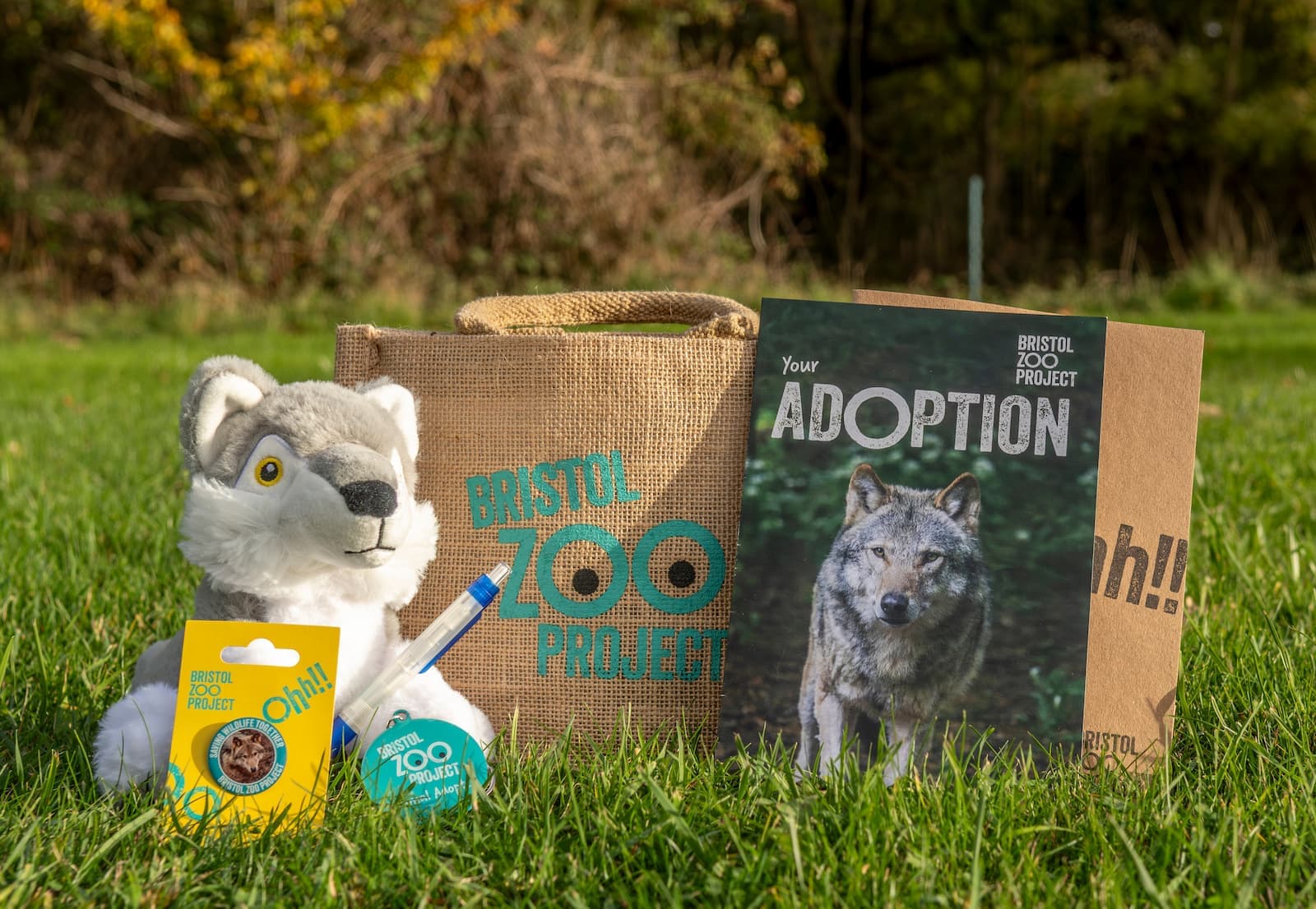 Adoption kit on grass with a plush wolf, pen, brochure featuring a wolf, and a burlap bag labeled "Bristol Zoo Project."